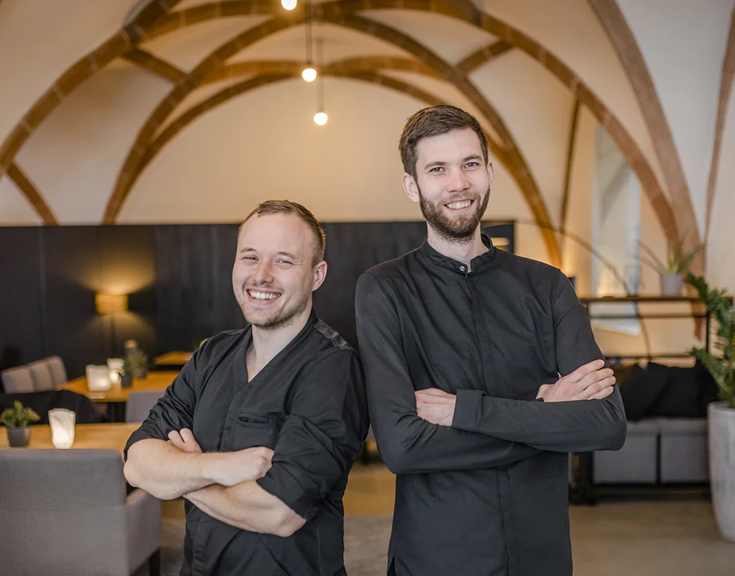 Zwei Köche im Innenraum des Restaurants zwölf87 in Ravensburg stehen nebeneinander, verschränken die Arme und lächeln in die Kamera