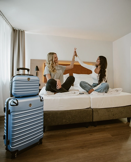 Zwei Frauen, eine blondhaarig und eine braunhaarig, sitzen lachend auf dem Bett in einem Hotelzimmer des Hotels zwölf87 – Traumwerk in Ravensburg und klatschen sich in die Hände
